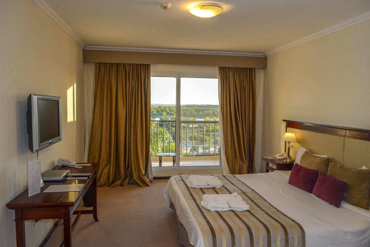A hotel room with a bed, a desk with a computer, a television, and a balcony with curtains.