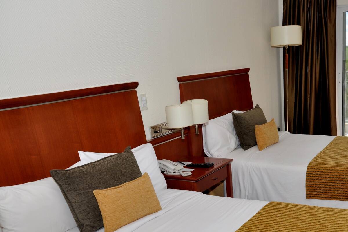 A hotel room with two beds, each with white linens and brown throw blankets. There is a nightstand with a lamp and a telephone.