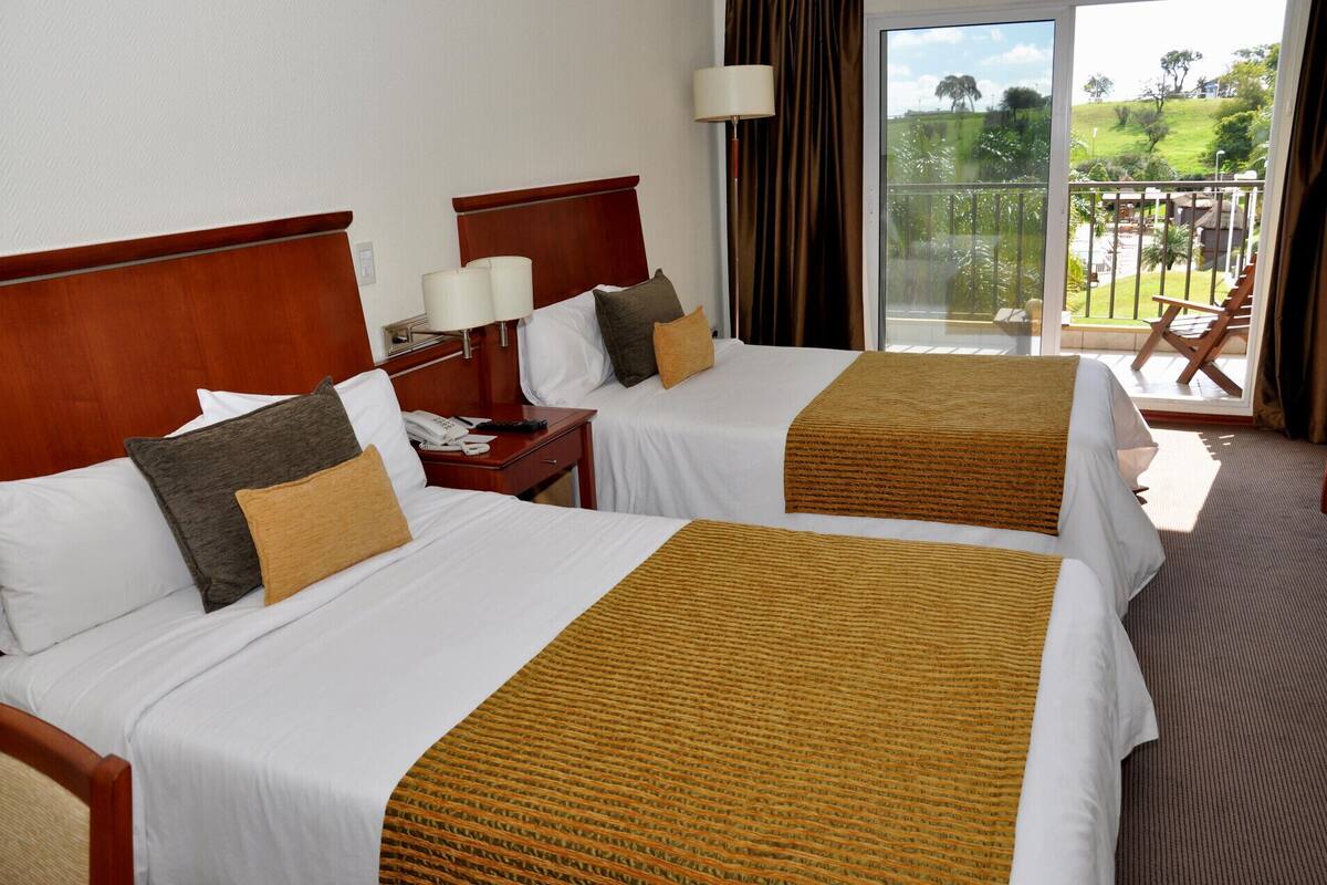 A hotel room with two beds, a nightstand, a lamp, and a view of the outdoors.