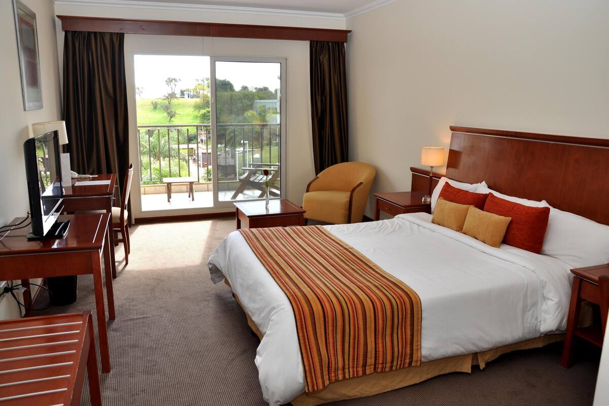 A hotel room with a large bed, a desk with a computer, a chair, a nightstand with a lamp, and a balcony with a table and chairs.