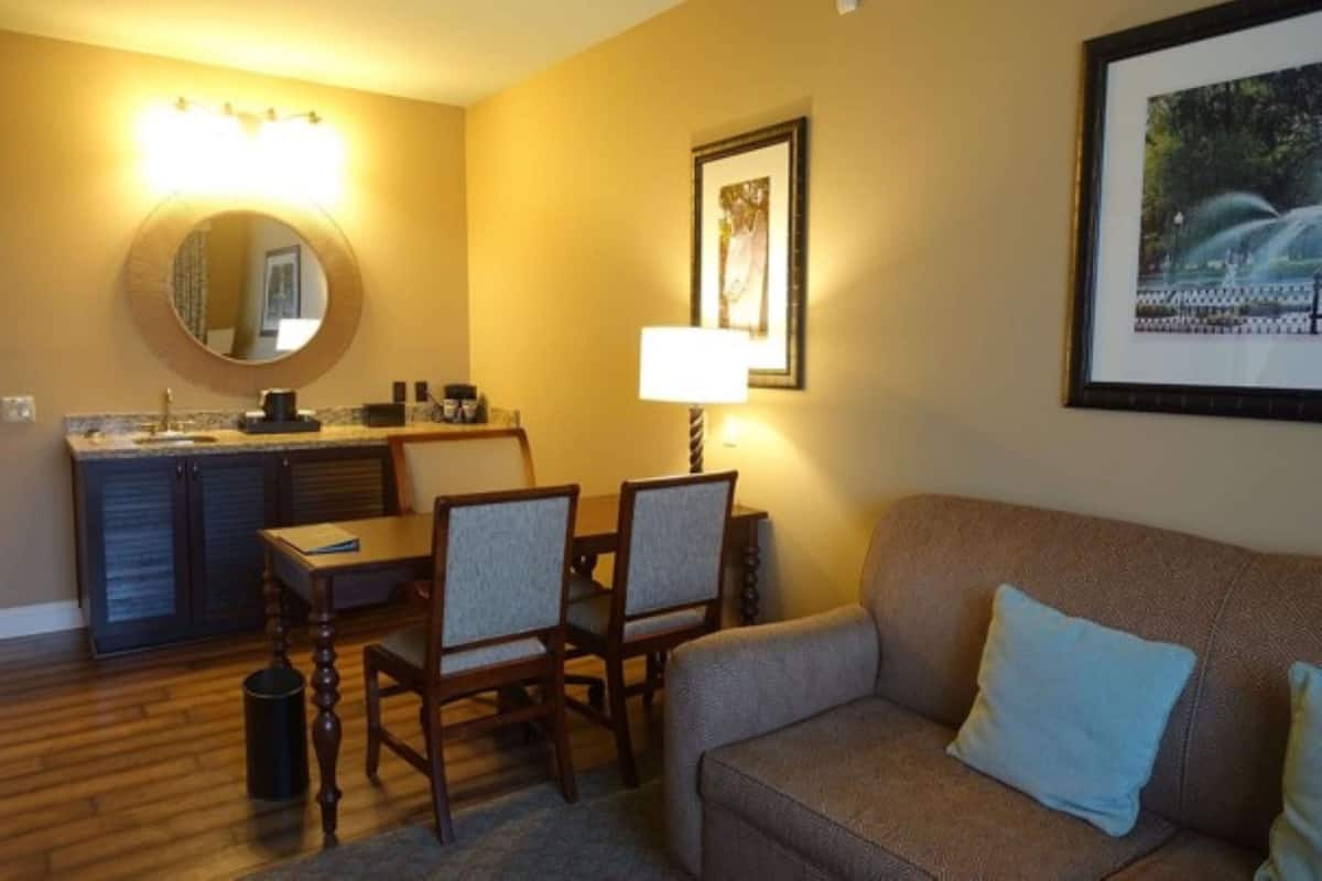 A hotel room with a dining area, a sofa, a framed picture on the wall, and a round mirror with lights above the sink.