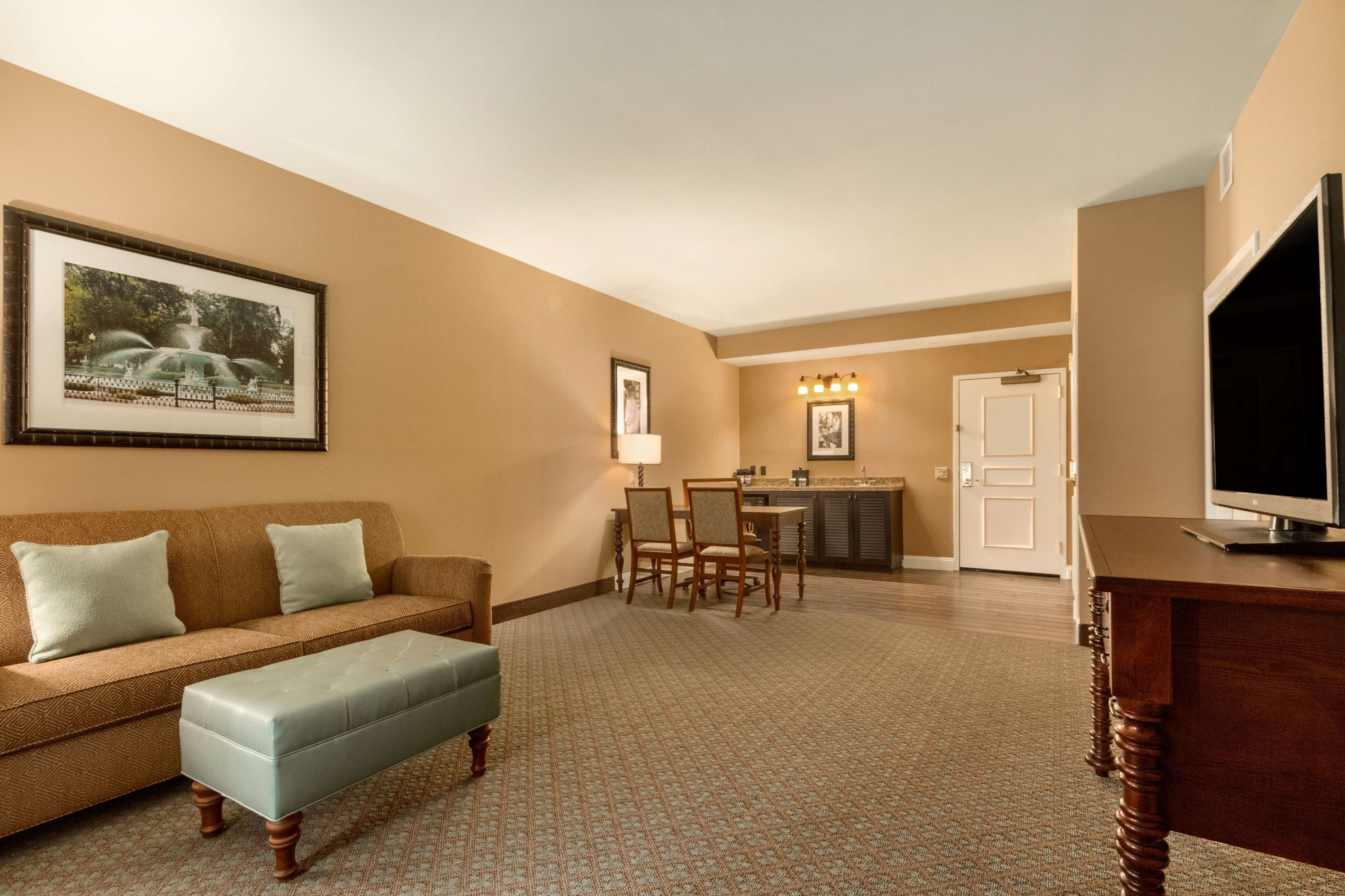 A hotel room with a brown sofa, a light blue ottoman, a dining table with chairs, a television on a wooden stand, and a framed picture on the wall.