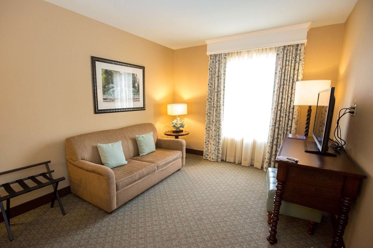 A hotel room with a beige sofa, a small table with a lamp, a desk with a mirror, and a framed picture on the wall.