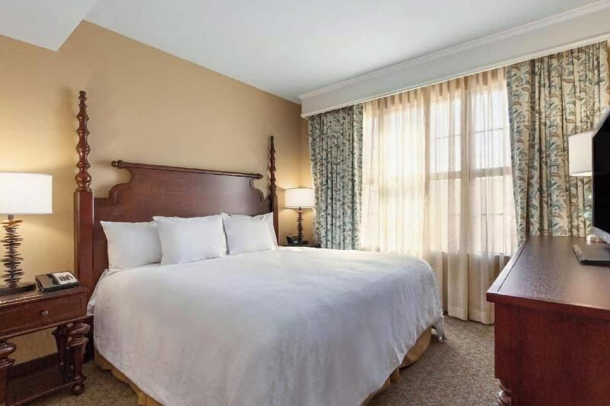 A hotel room with a large bed, two bedside tables with lamps, a desk, and a window with curtains.