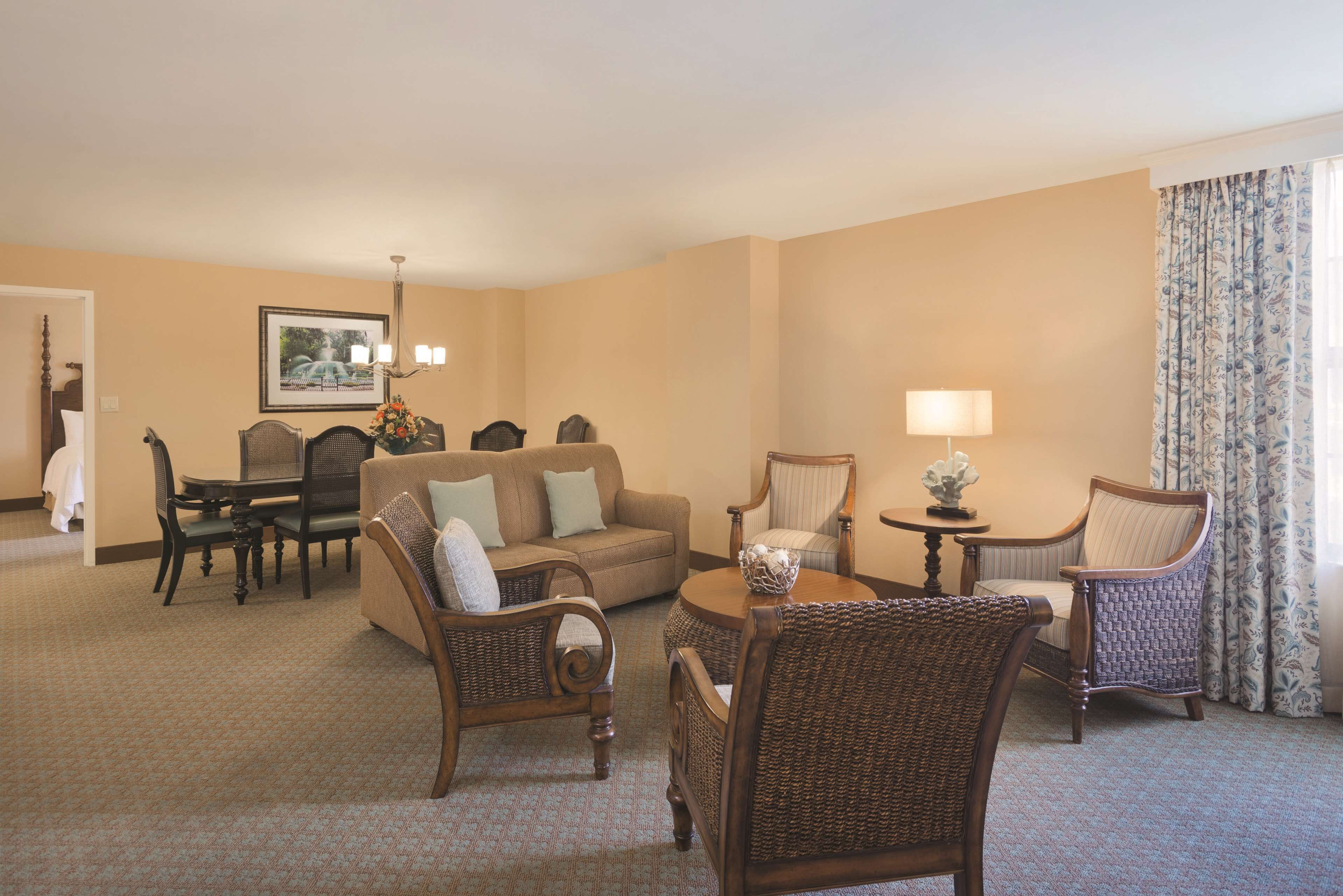 A hotel room with a beige carpet, a sofa, two wicker chairs, a dining table with chairs, a lamp, and a framed picture on the wall.