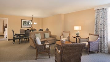 A hotel room with a beige carpet, a sofa, two wicker chairs, a dining table with chairs, a lamp, and a framed picture on the wall.