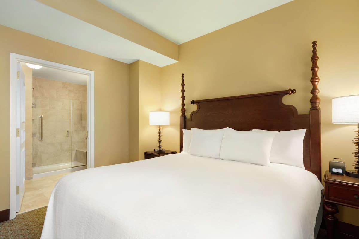 A hotel room with a large bed, a wooden headboard, a nightstand with a lamp, and a glass shower enclosure.