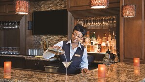 A bar area with a bartender pouring a drink into a martini glass, a television mounted on the wall, and a variety of bottles and glasses on the shelves. - Embassy Suites by Hilton Savannah Historic District (Savannah)