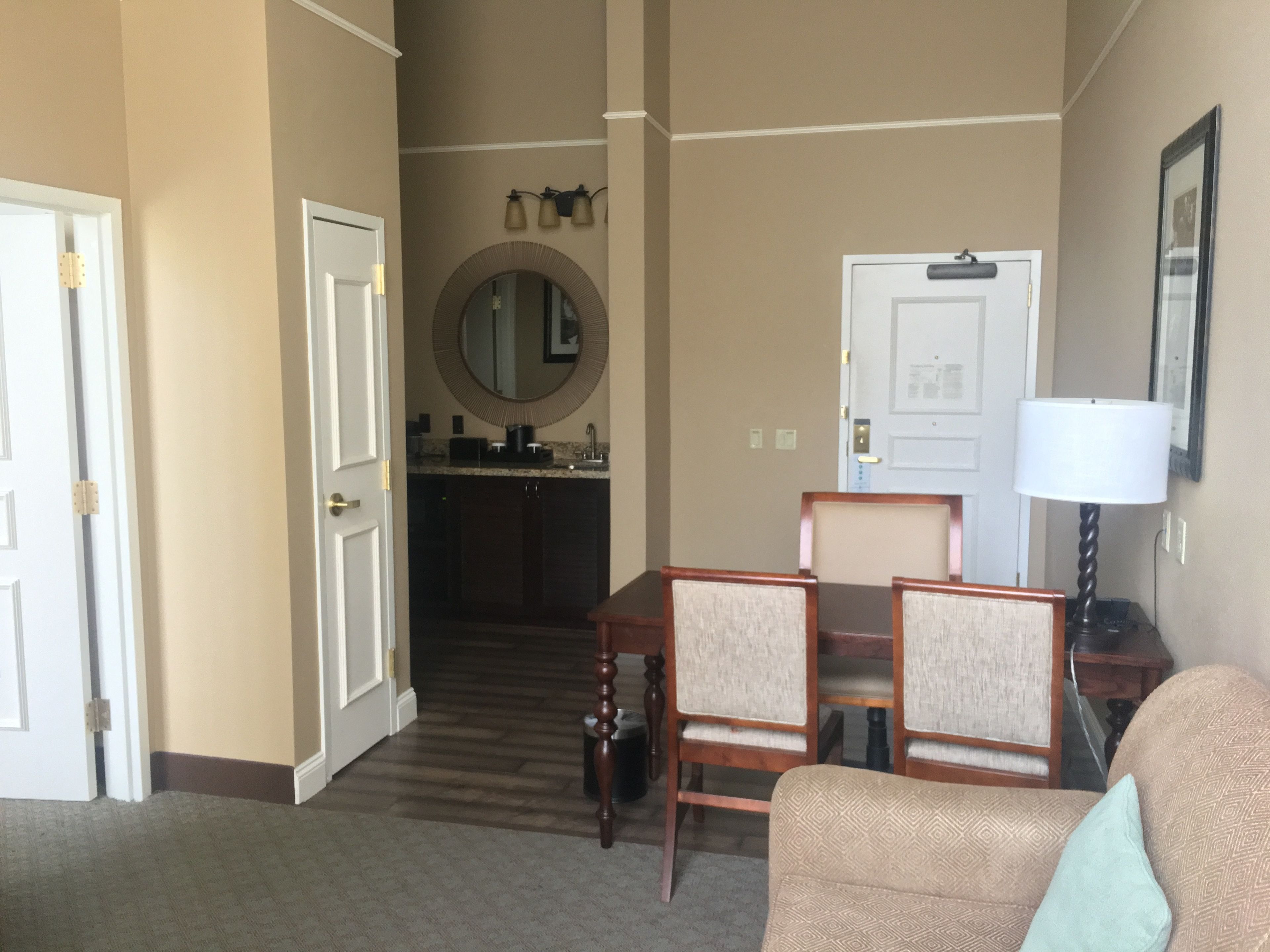 Um lobby de hotel com uma mesa de recepção, dois poltronas, uma mesa de jantar, um espelho e uma porta que dá para uma cozinha.