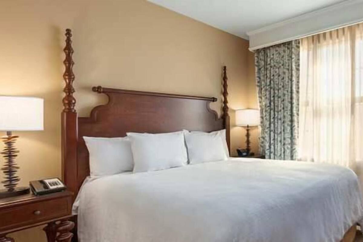 A hotel room with a large bed, two bedside tables, a desk, and a window with curtains.
