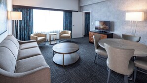 Una habitación de hotel moderna con un sofá, dos sillones, una mesa de centro redonda, una mesa de comedor con sillas, un televisor y una lámpara.