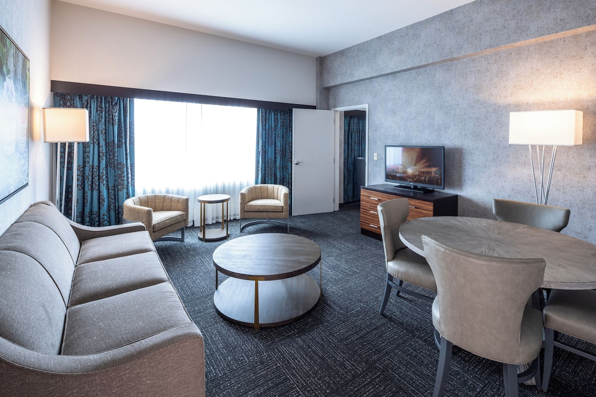 A modern hotel room with a sofa, two armchairs, a round coffee table, a dining table with chairs, a TV, and a lamp.