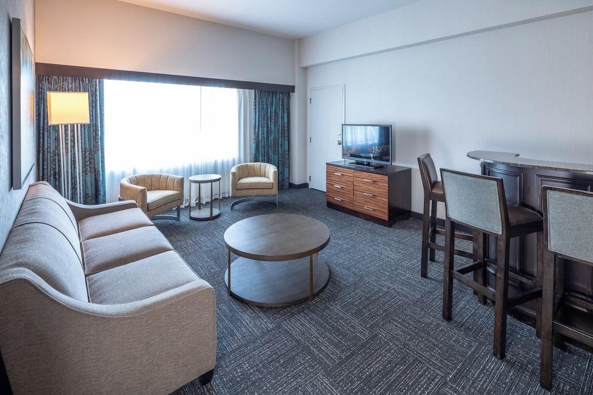 A hotel room with a sofa, armchair, round coffee table, TV, and a small bar area.