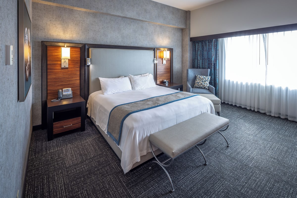 A hotel room with a large bed, bedside tables with lamps, a chair, and a bench.