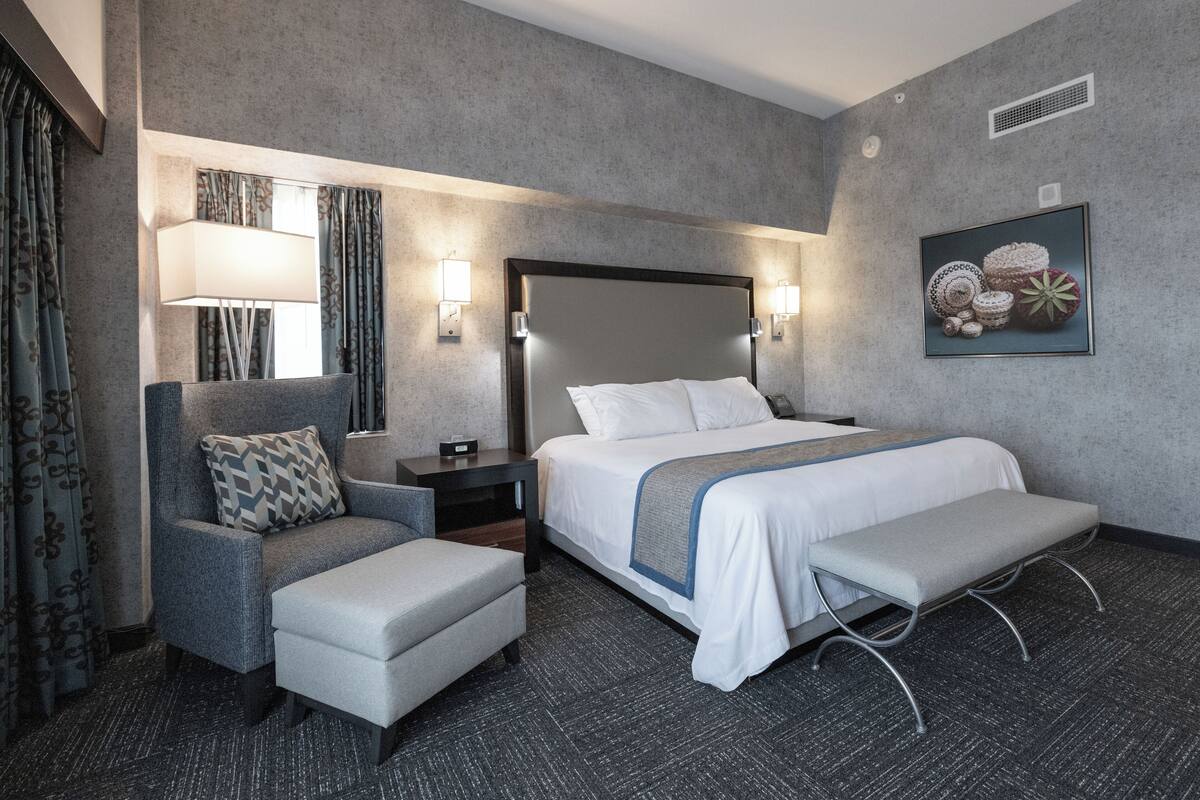 A hotel room with a bed, a chair, a bench, a nightstand, a lamp, and a wall-mounted picture.