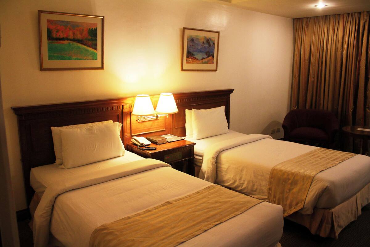 Una habitación de hotel con dos camas, una mesita de noche con lámpara y dos cuadros en la pared.