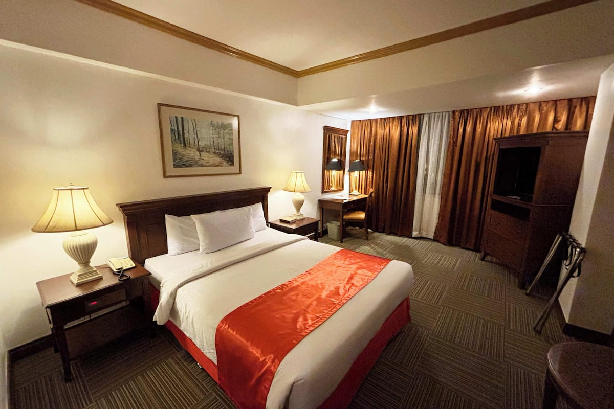 Una habitación de hotel con una cama grande, dos mesitas de noche con lámparas, un escritorio con una silla, un televisor y un cuadro en la pared.