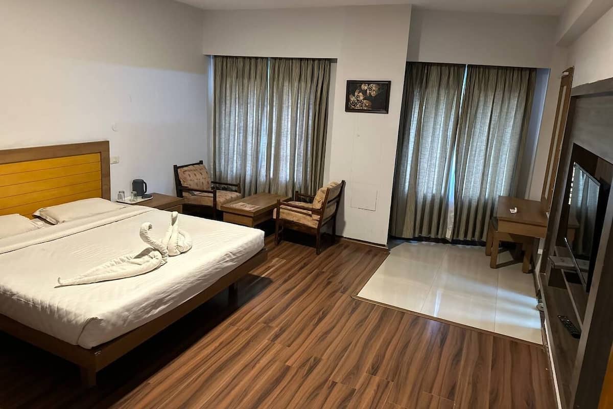 A hotel room with a wooden floor, a bed with a white comforter, two chairs, a small table, a television, and curtains.