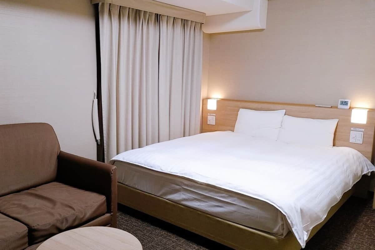 Une chambre d’hôtel avec un lit, un canapé, une fenêtre avec des rideaux et un luminaire fixé au mur.