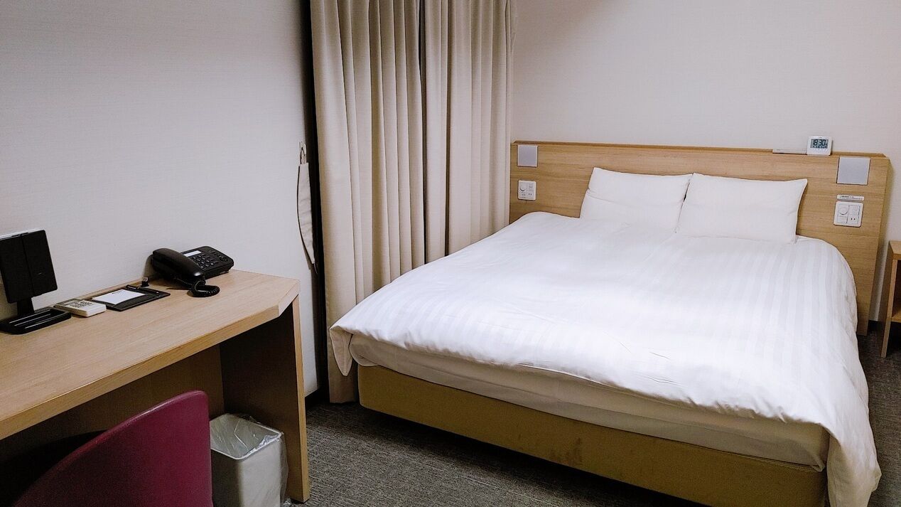 A hotel room with a bed, a desk with a telephone, a chair, and a trash bin.