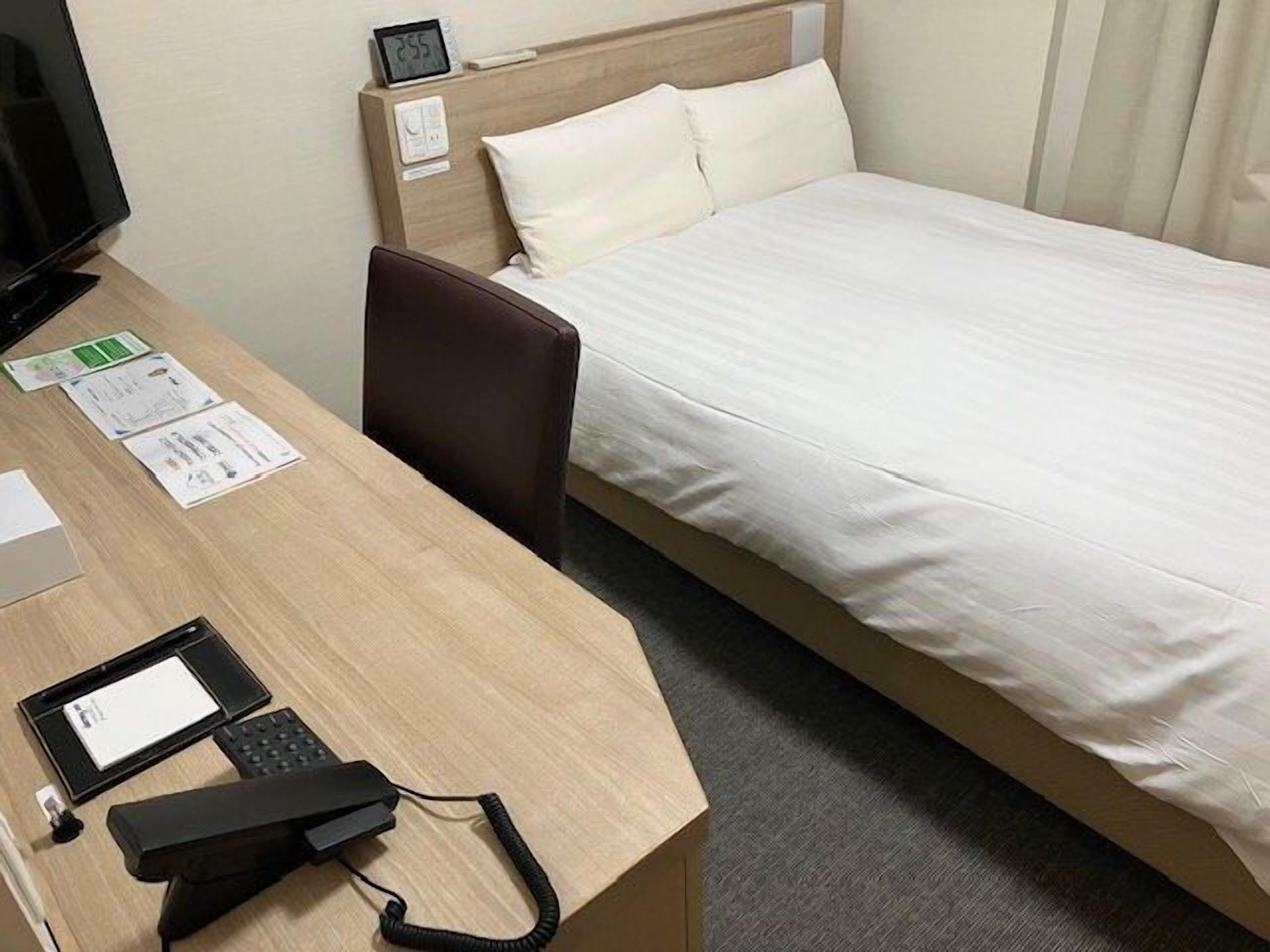A hotel room with a bed, a desk with a computer, a chair, and a bedside table with a clock.