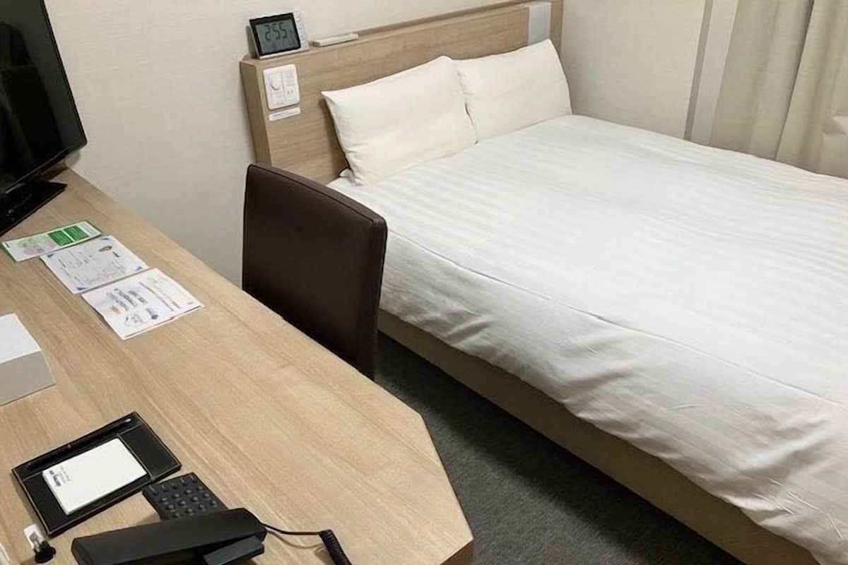 A hotel room with a bed, a desk with a computer, a chair, and a bedside table with a clock.