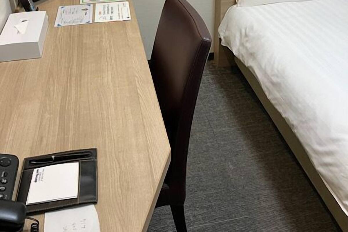 A hotel room with a wooden desk, a television, a bed with white bedding, a nightstand with a clock, and a chair.