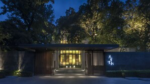 Una entrada de edificio moderna con una galería techada, iluminada por una luz cálida, rodeada de árboles.