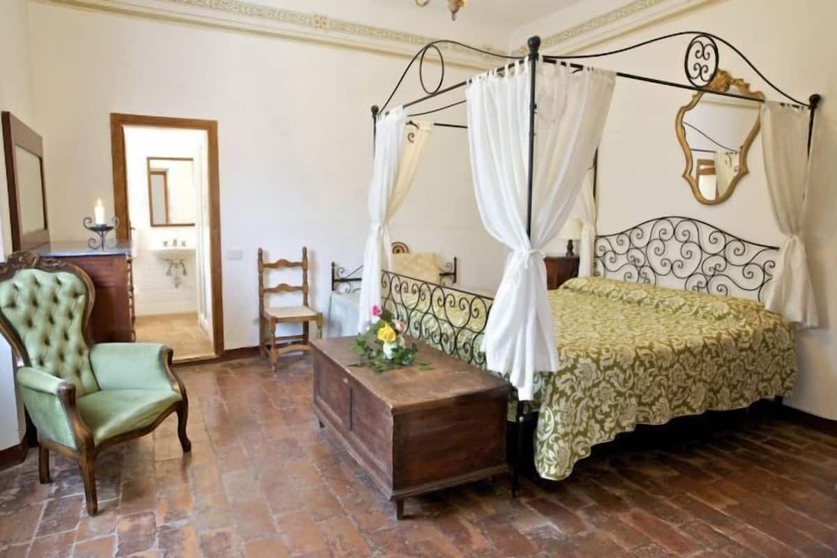 Un dormitorio con cama con dosel, una mesita de noche de madera, un sillón verde y un banco de madera.