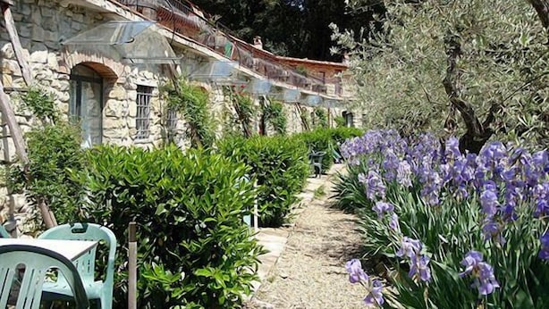 Un edificio de piedra con balcón, rodeado de vegetación y flores moradas.