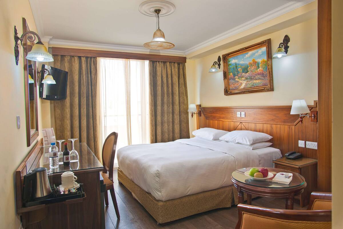 Une chambre d’hôtel avec un lit, un bureau doté d’une machine à café, une chaise, une petite table avec des fruits et un tableau au mur.