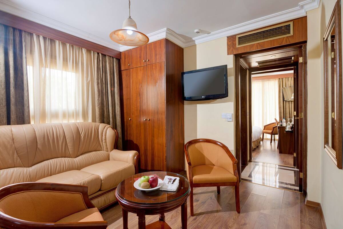Une chambre d’hôtel avec un canapé, des fauteuils, une table avec des fruits, une télévision et une porte donnant sur une salle de bain.