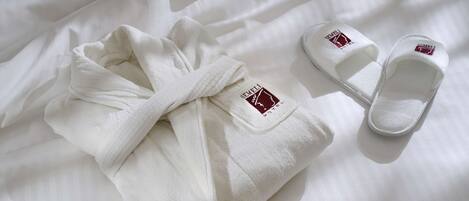 A neatly folded white bathrobe and a pair of white slippers with a logo on a bed.