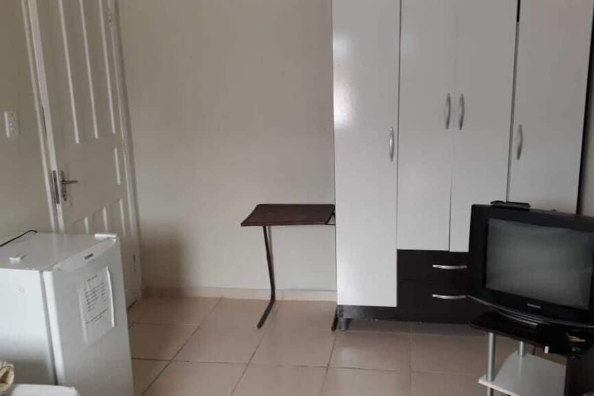 A room with a white wardrobe, a small table, a television on a stand, and a washing machine.