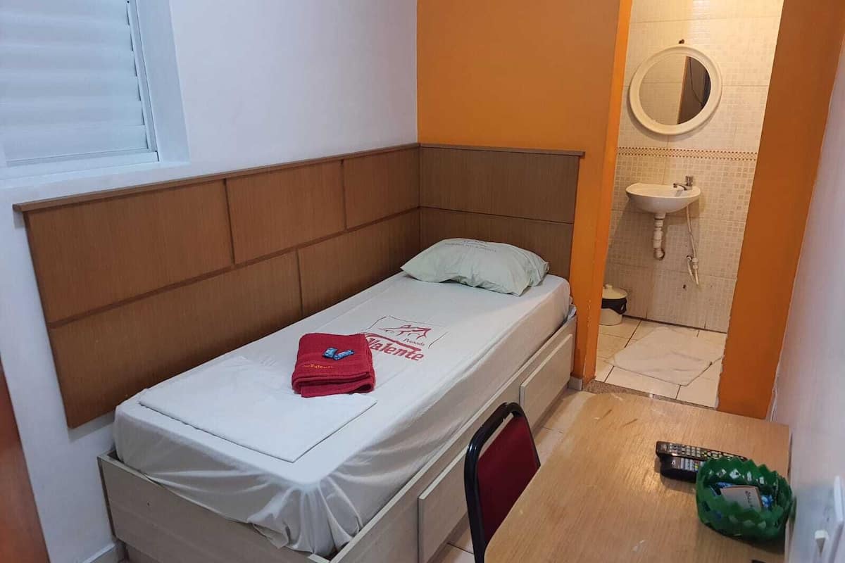 A small, single-bed room with a wooden headboard, a white bedspread, a red towel, a green basket, and a remote control on the floor.