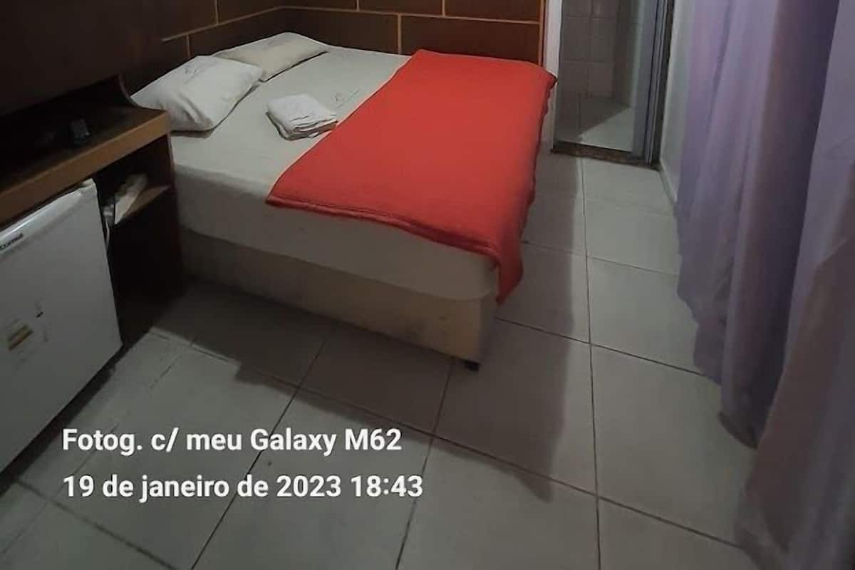 A small, simple room with a bed, a desk, a TV mounted on the wall, and a shower area.