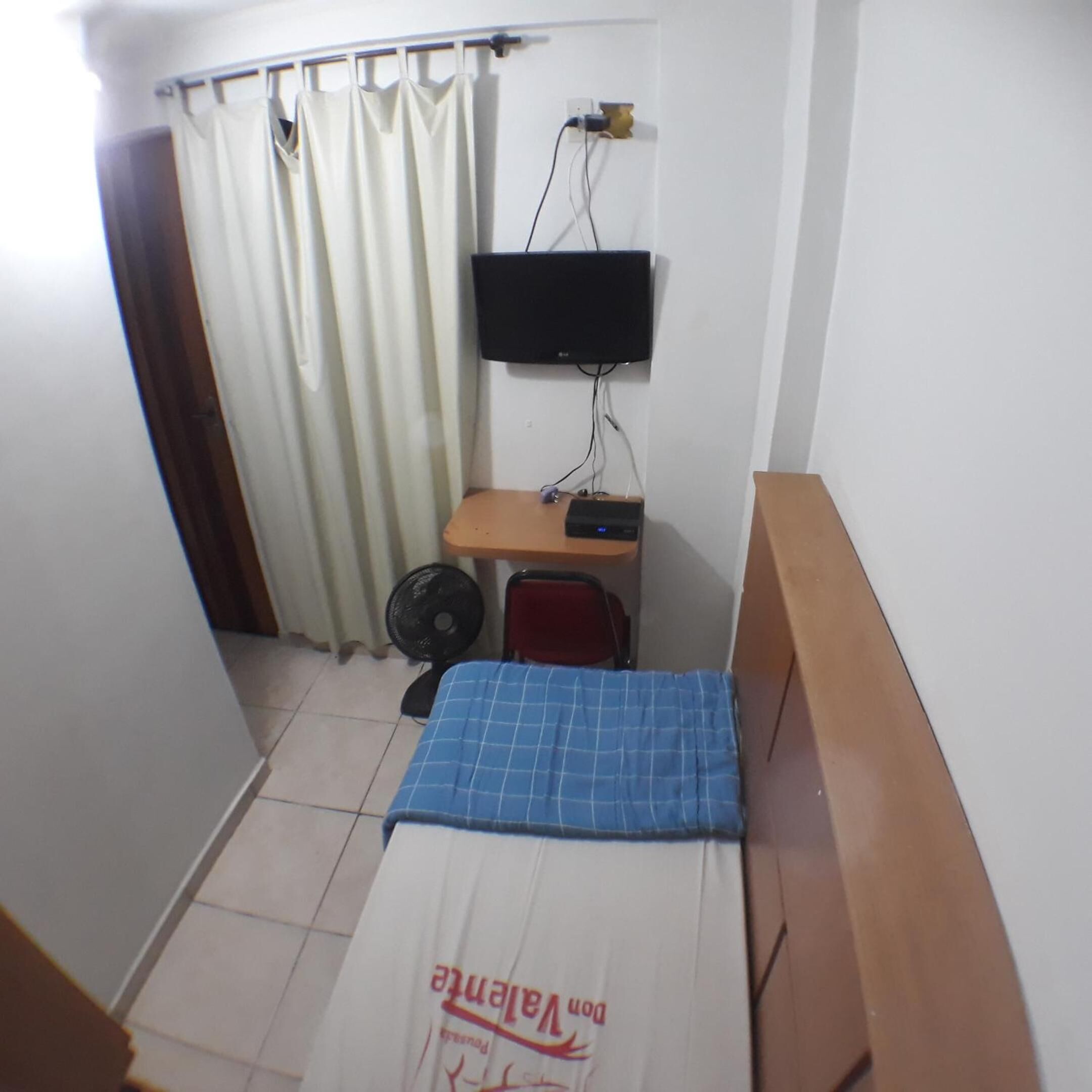 Une petite pièce comprenant un lit, un bureau, une chaise, un téléviseur et un ventilateur.