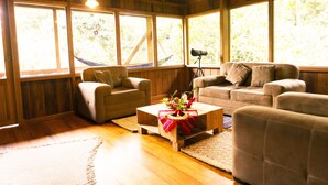 A cozy living room with wooden walls and large windows, a sofa, a chair, and a coffee table with a decorative centerpiece. - Casa Divina Eco Lodge (Mindo)