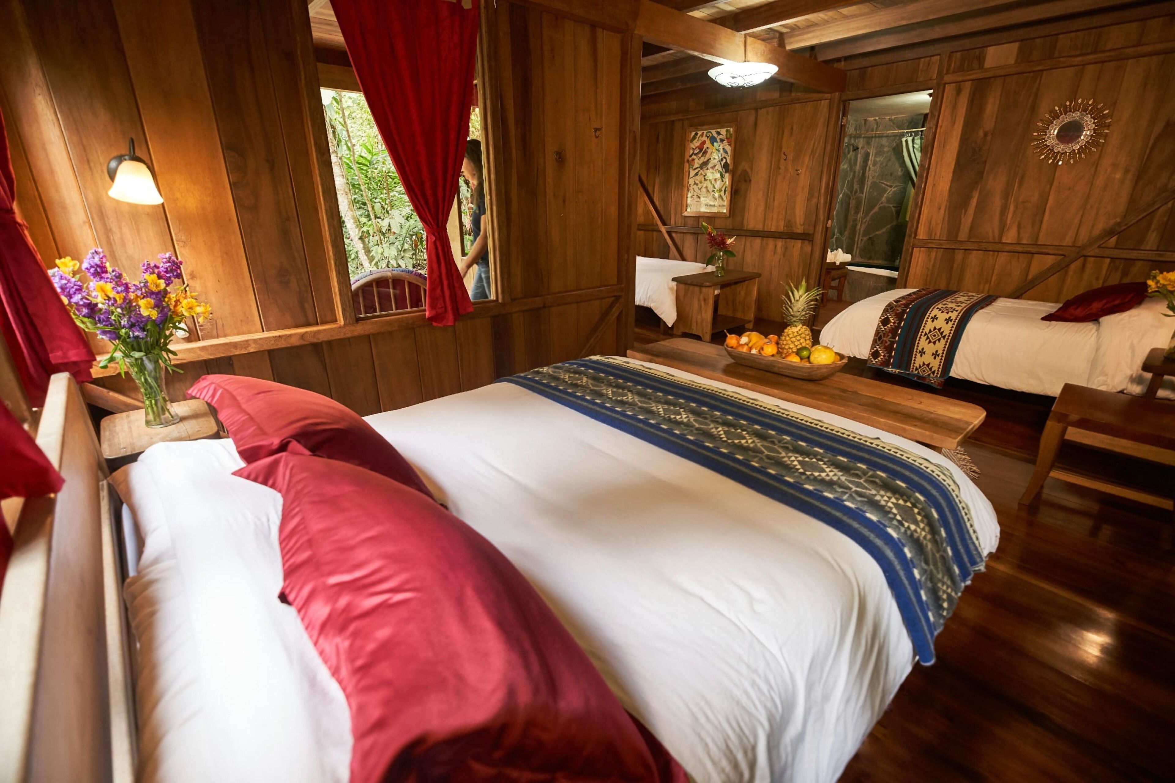 Quarto de cabana de madeira com uma cama, uma mesa pequena com frutas, um vaso de flores e uma janela com cortinas.