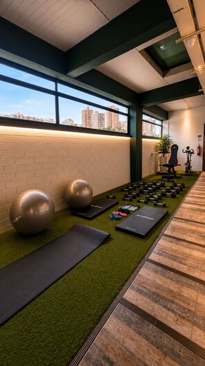 A gym with exercise equipment, a green exercise mat, and a view of buildings through large windows. - Hotel Provenza Garden (Medellín)