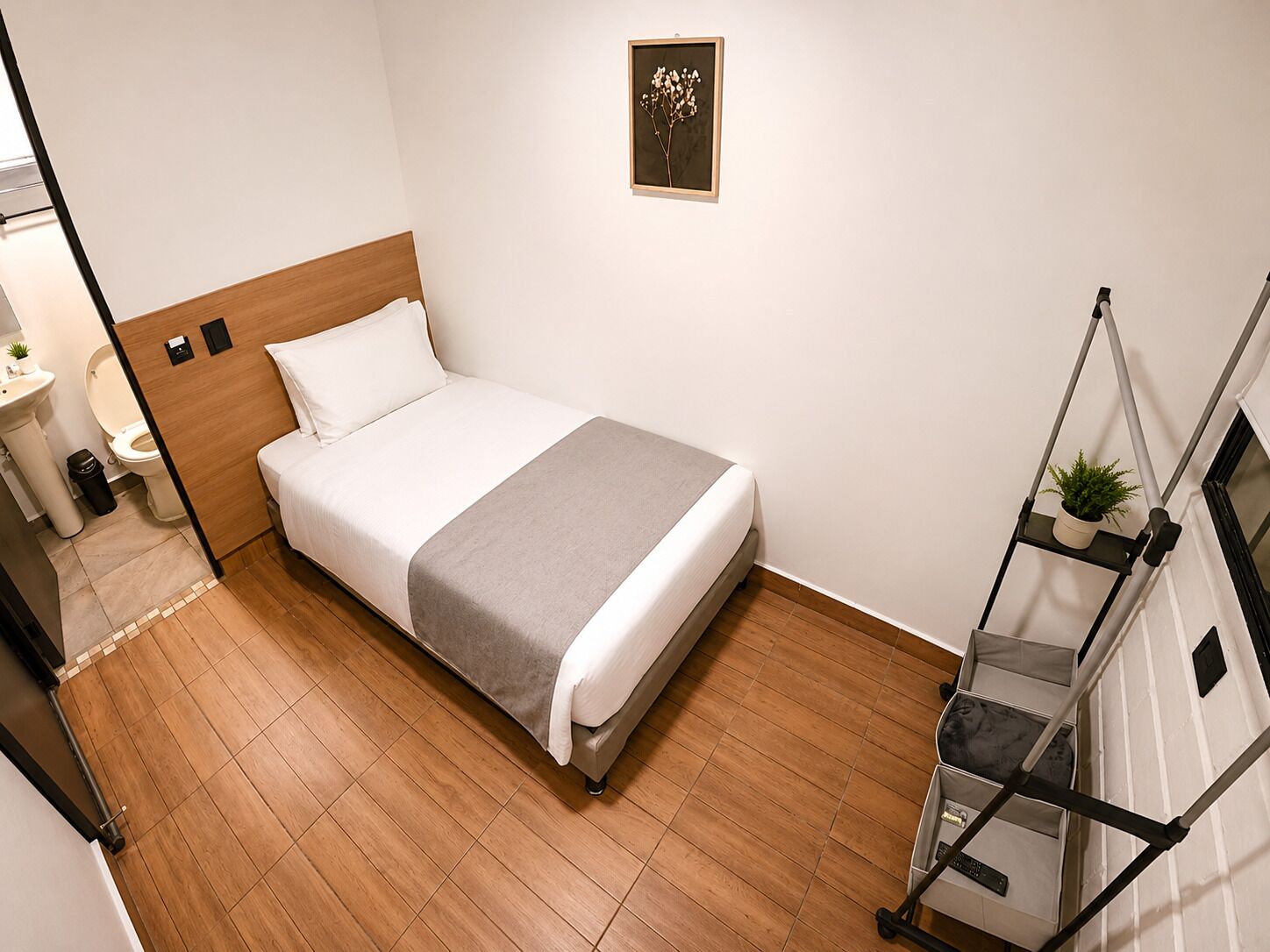 Habitación de hotel ordenada con cama, mesita de noche, una pequeña planta en maceta y un cuadro colgado en la pared.