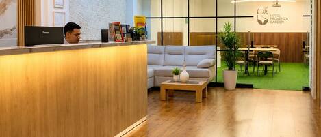 A hotel reception area with a wooden reception desk, a sofa, a coffee table, and a dining area with a green carpet.