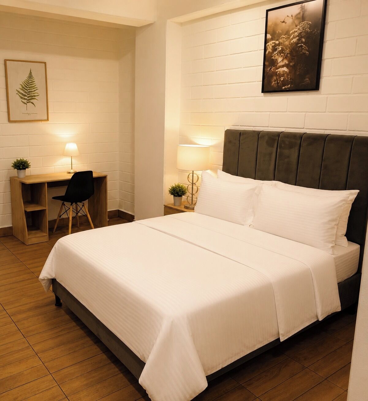 A neatly made bed with white bedding, a wooden desk with a chair, a nightstand with a lamp, and a framed botanical print on the wall.