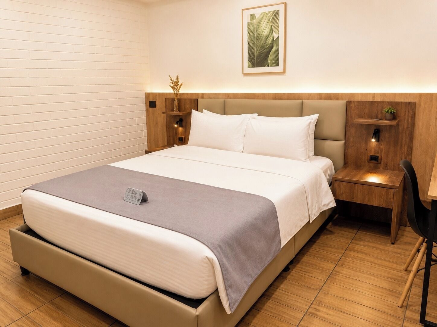 Quarto de hotel com uma cama grande, uma mesa de cabeceira, uma cadeira, uma pequena escrivaninha e uma estante fixada na parede.