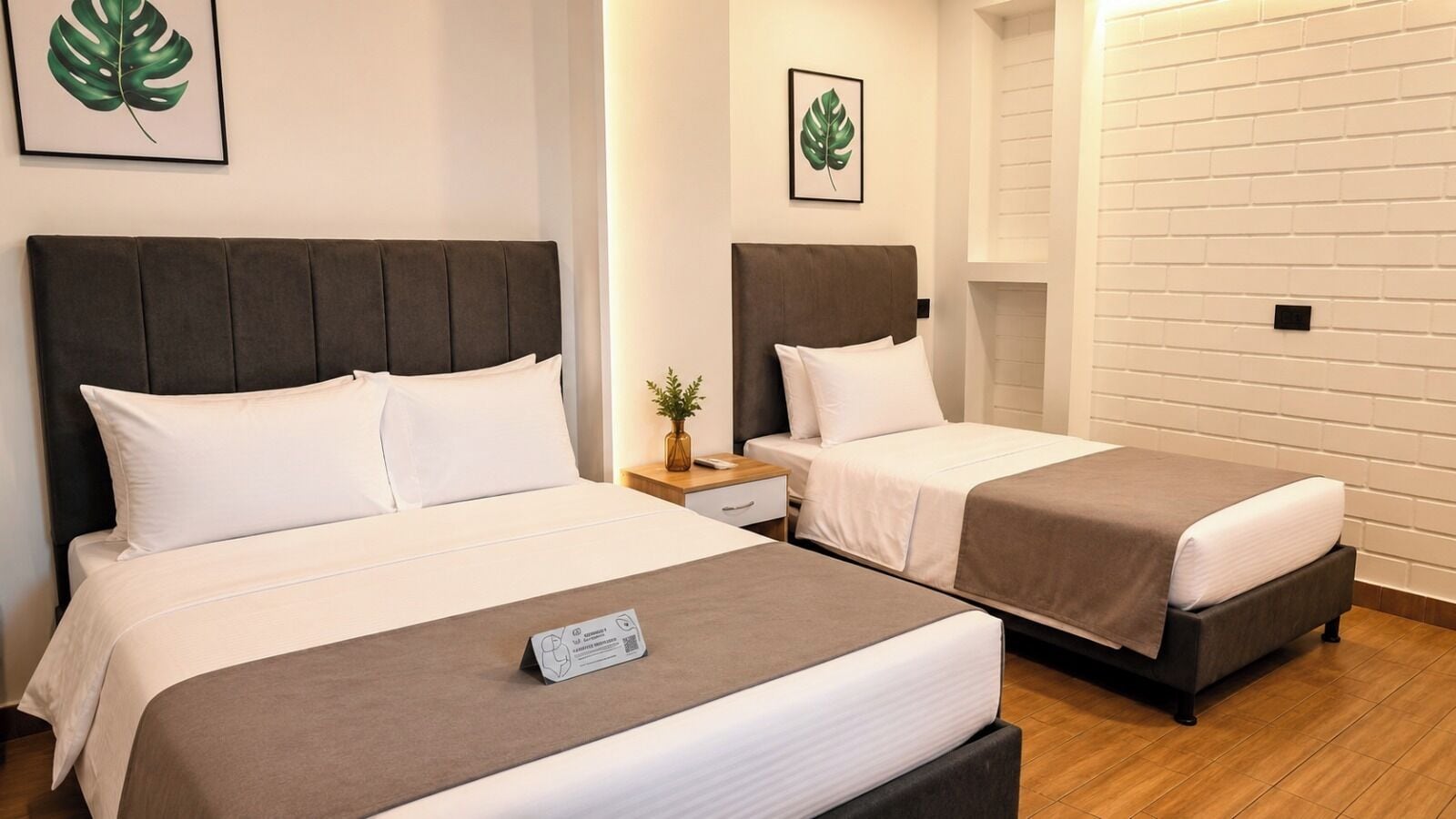 A hotel room with two beds, a bedside table with a lamp, and a wall-mounted light fixture.