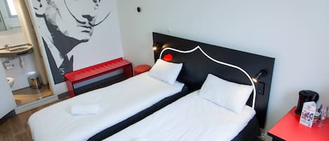 A hotel room with two beds, a red table, a wall-mounted light, and a bathroom visible through an open door.