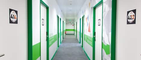 Un lungo corridoio stretto con porte a cornice verde, targhe con numeri e decorazioni geometriche alle pareti.