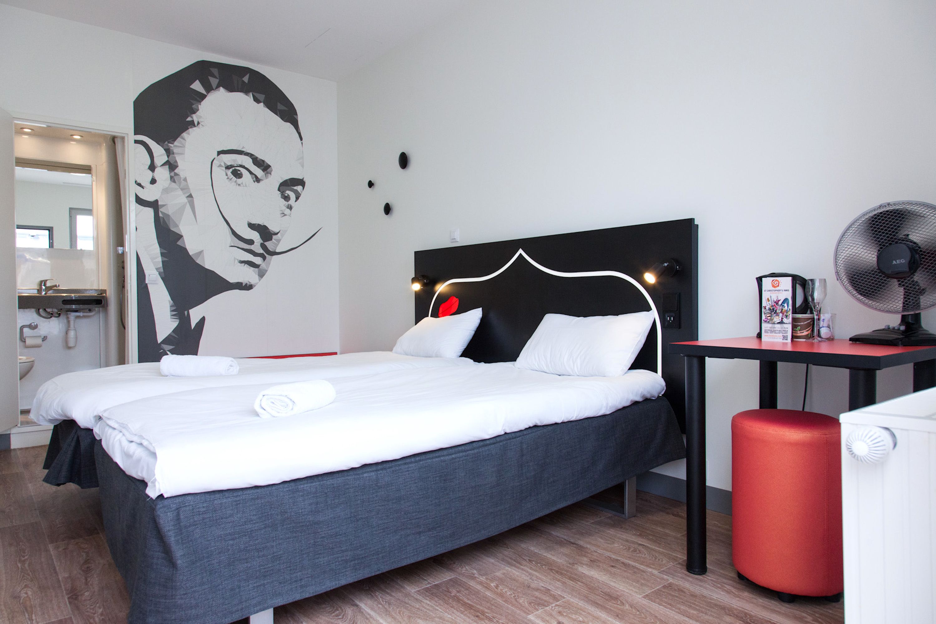 Una habitación de hotel moderna con una cama grande, un escritorio, un reposapiés rojo y un ventilador fijado a la pared.
