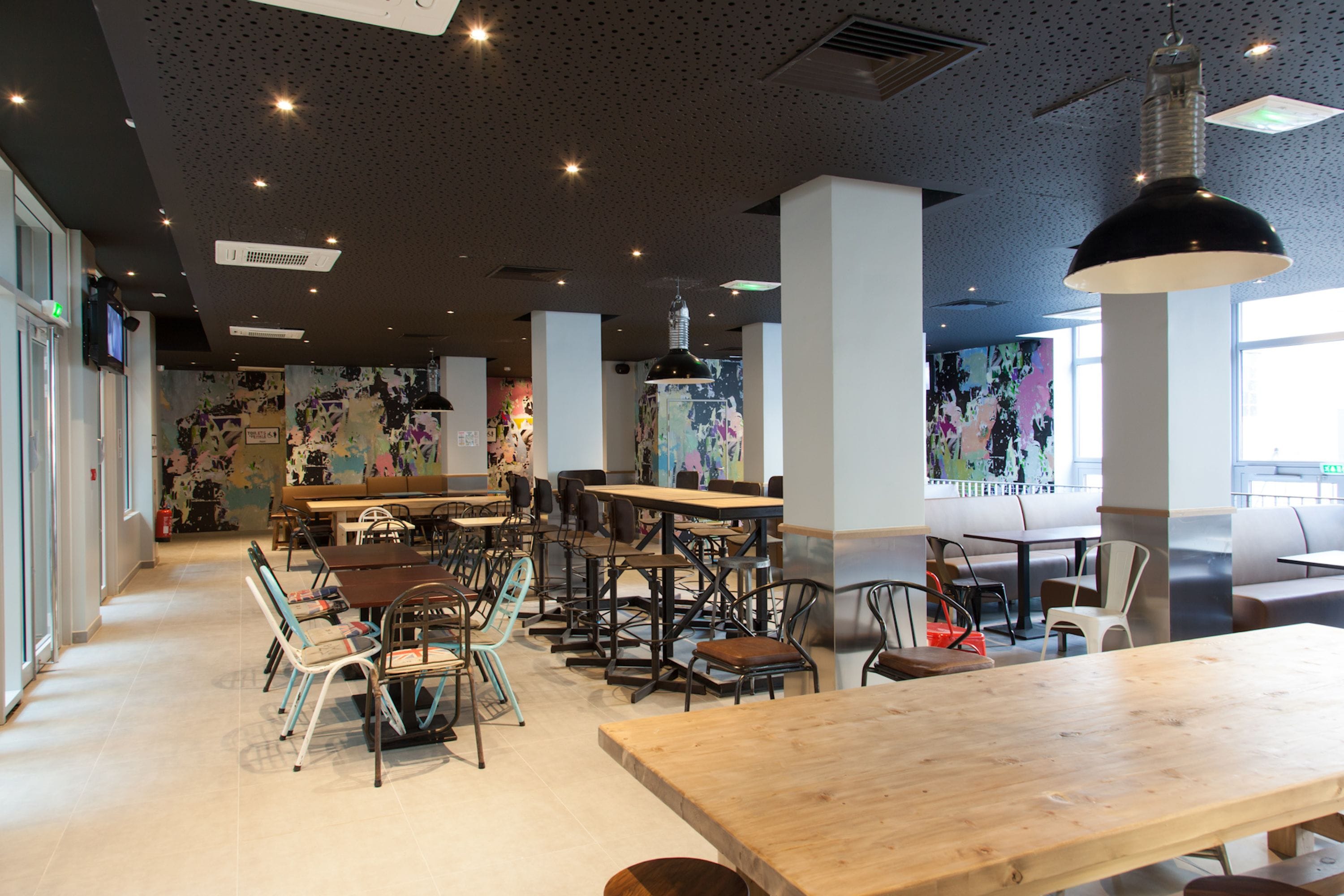 Un intérieur de restaurant moderne avec des tables et des chaises hautes, un grand luminaire suspendu et une fresque colorée sur le mur.