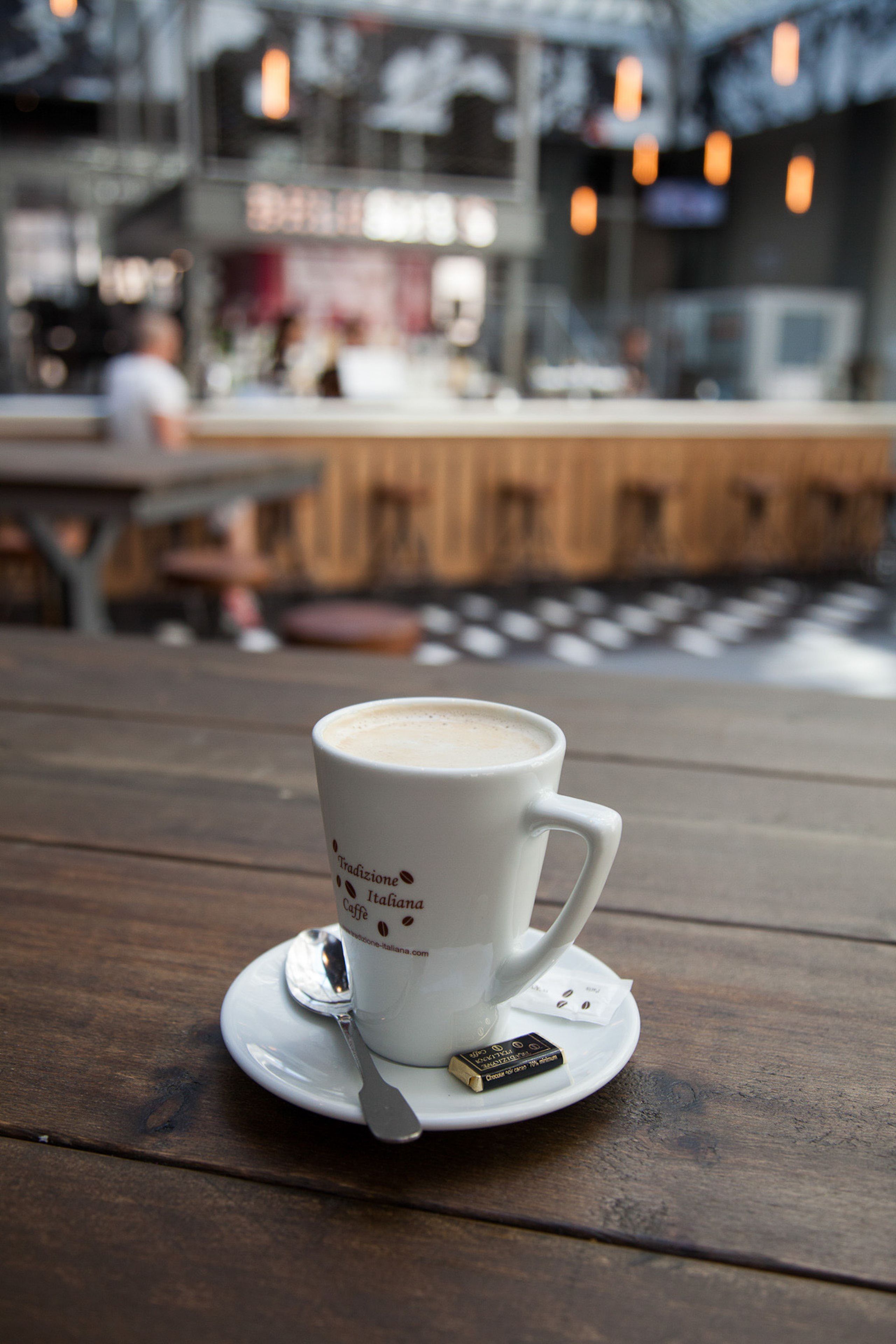 A cup of coffee on a wooden table with a spoon and a small packet, in a cafe with a blurred background.